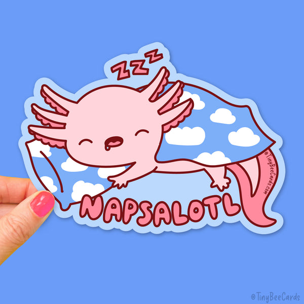 A hand holds a die-cut sticker featuring a sleeping pink axolotl under a blue cloud-patterned blanket, with the word 'NAPSALOTL' in pink letters. The sticker is shown against a solid blue background.
