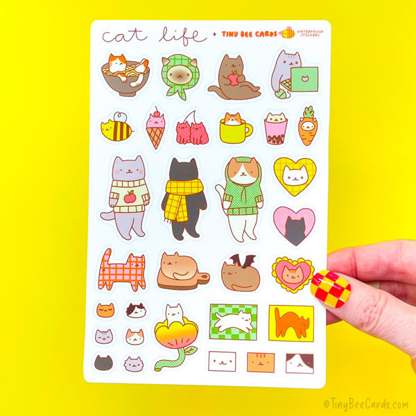Cat Life Vinyl Sticker Sheet. A hand holding a 4x6 sticker cat life vinyl sticker sheet. Stickers on it include cats at work, cats on their day off, cats wearing sweaters, cherry cats, ice cream cats, cute cat faces, loaf cats, cats emerging from tulips and more. Variety of different cats including calico, black cats, siamese cats, orange cats, grey cats, striped cats, and white cats.