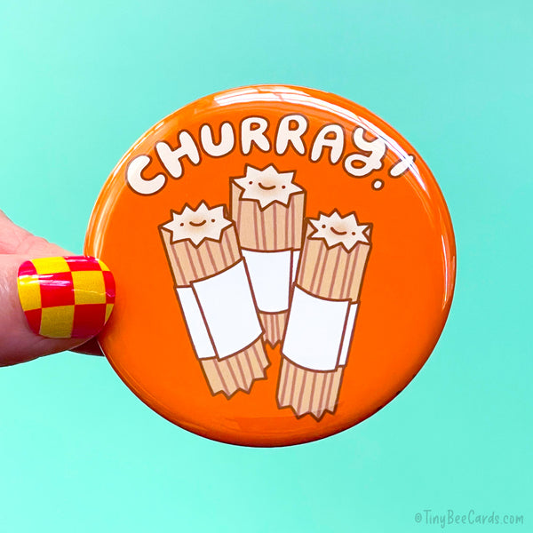 A button badge with a hand drawn illustration of 3 churro with happy faces and the hand drawn text "Churray!" on an orange background. 