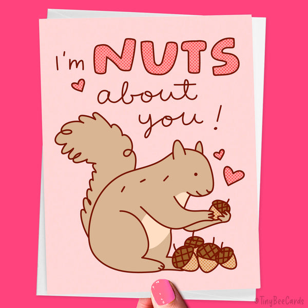 Valentine's Day card with a cute hand drawn squirrel holding a nut and text 'I'm NUTS about you!' on a pink background with nuts and hearts around.