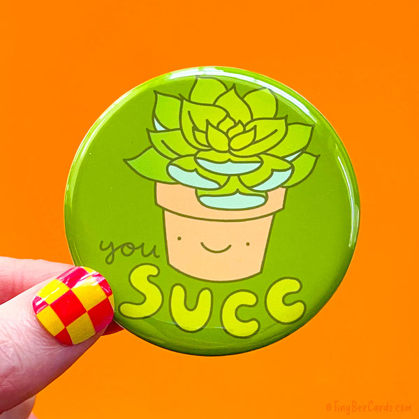 A button badge with a hand drawn illustration of a cute succulent plant in a terracotta pot with a smiley face, and the text "You Succ."