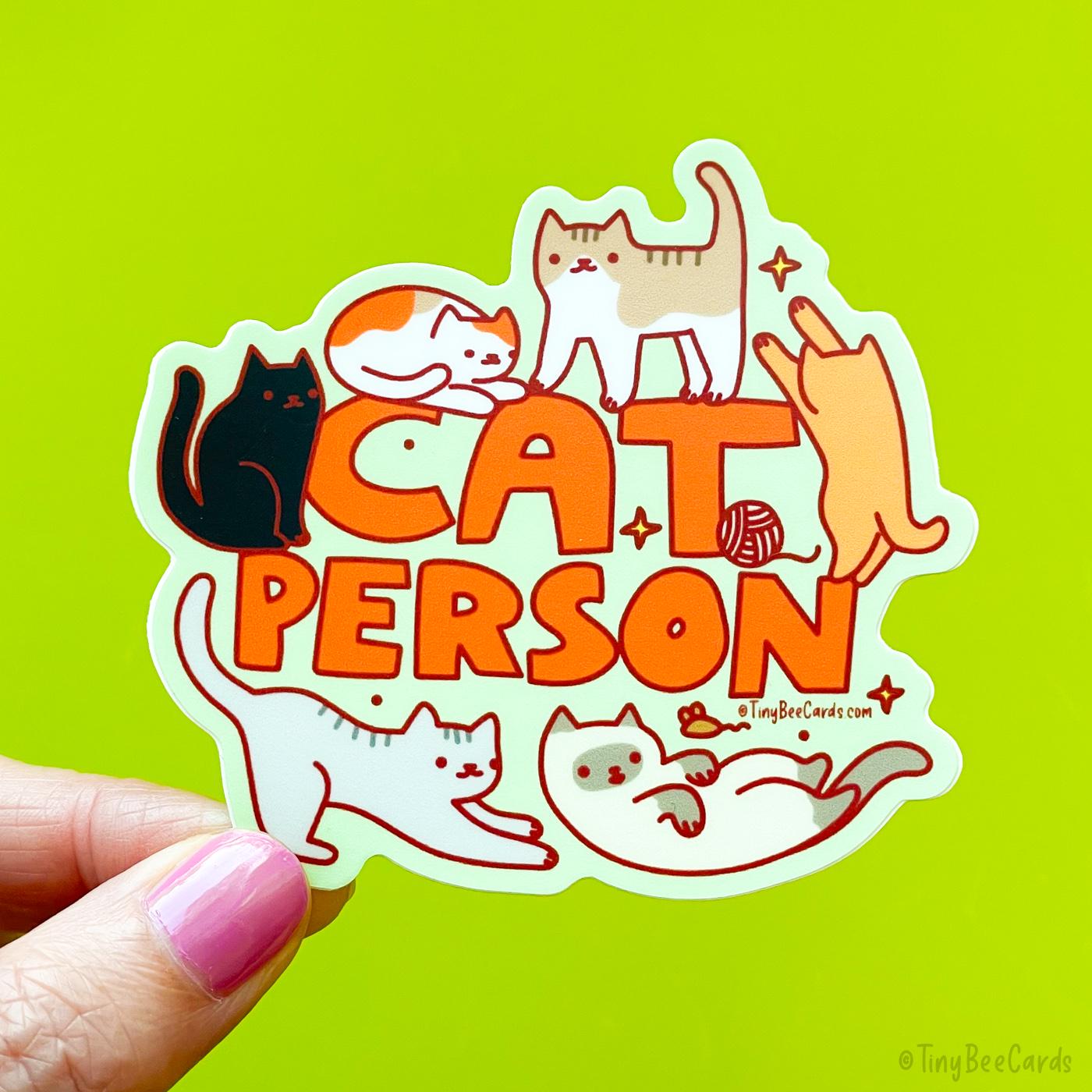 A die cut sticker that says &quot;cat person&quot; with 6 cats surrounding the text, including a calico, a black cat, a brown and white striped cat, an orange cat, a siamese, and a grey striped cat. They are playing, sitting and standing.