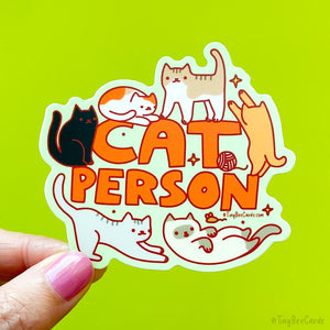 A die cut sticker that says &quot;cat person&quot; with 6 cats surrounding the text, including a calico, a black cat, a brown and white striped cat, an orange cat, a siamese, and a grey striped cat. They are playing, sitting and standing.