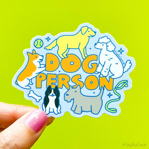 a die cut vinyl sticker that says &quot;dog person&quot; in hand drawn letters, with 5 dogs surrounding the text, including a golden retriever, a white poodle, a corgi, a terrier and a frenchie. They are playing and standing, with toys and bones.