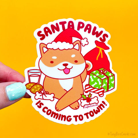A die cut vinyl sticker featuring a cute shiba inu dog surrounded by gifts, dog bones and chew toys, and wearing a santa hat. Pun text says "Santa Paws is coming to town!"