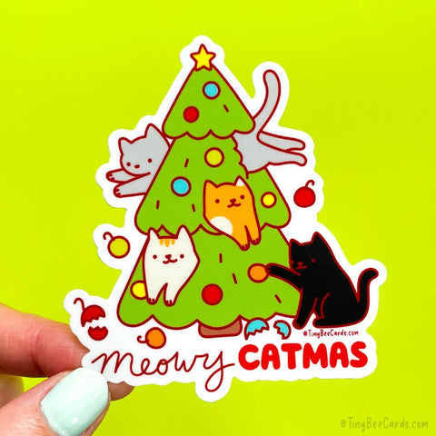 A die cut vinyl sticker featuring 4 cats (a black cat, a calico, an orange cat, and a grey cat) jumping into and being mischievous with the holiday christmas tree and breaking a few ornaments.