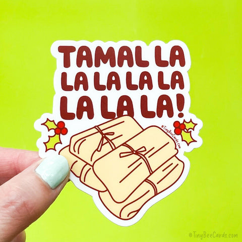 A Christmas holiday die cut vinyl sticker featuring a pile of delicious looking tamales with the festive holiday pun "Tamal la la la la la la la" like the fa la la holiday song. Two holly plants decorate the sticker. 