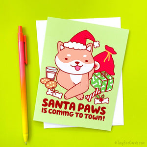 A cute shiba inu dressed as santa with cookies, milk, holiday dog bones, and presents on a Christmas card with the pun "Santa Paws is Coming to Town!"